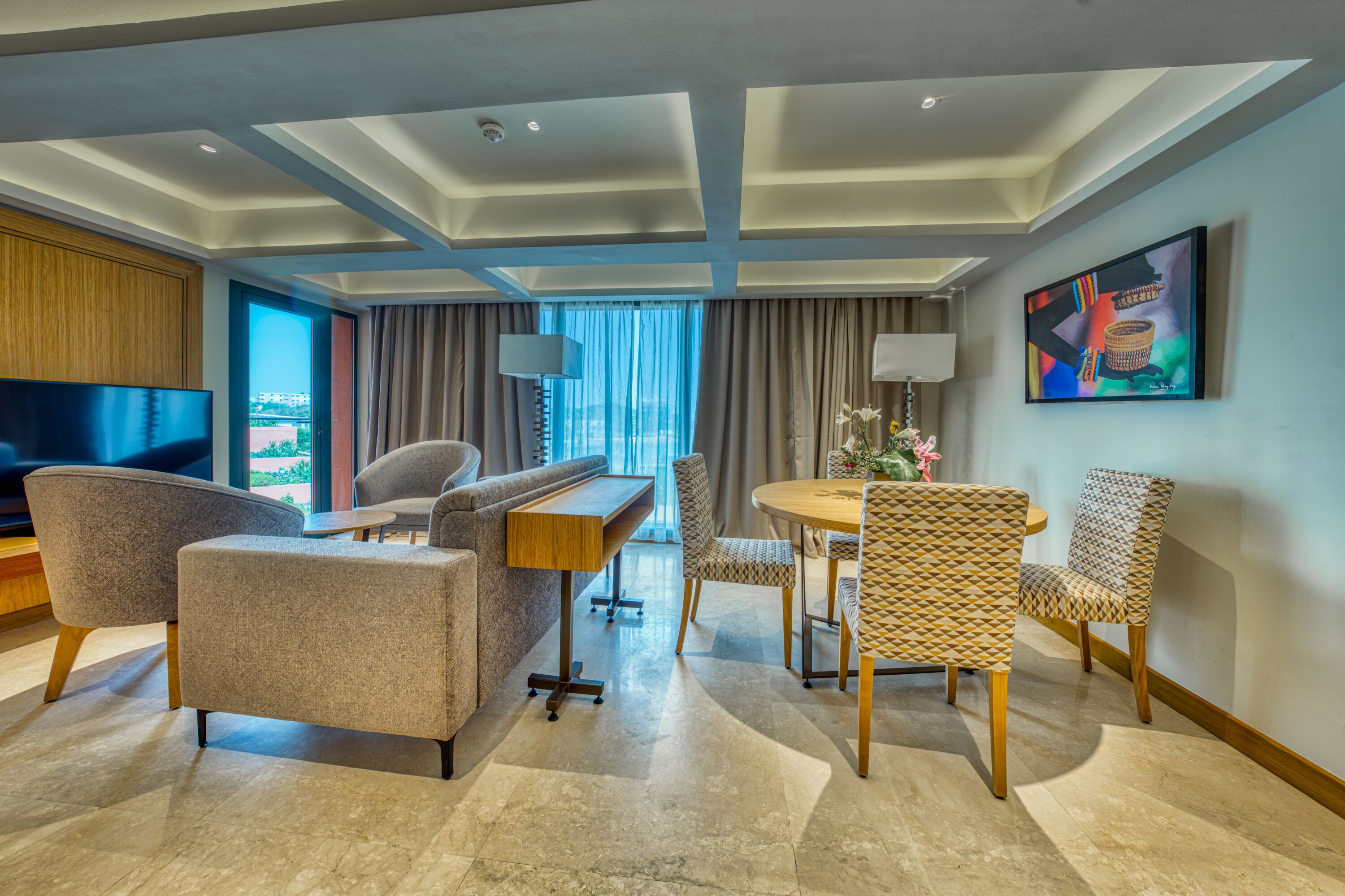 A modern hotel room with a flat-screen TV, comfortable seating, a dining area, and a large window offering a city view.
