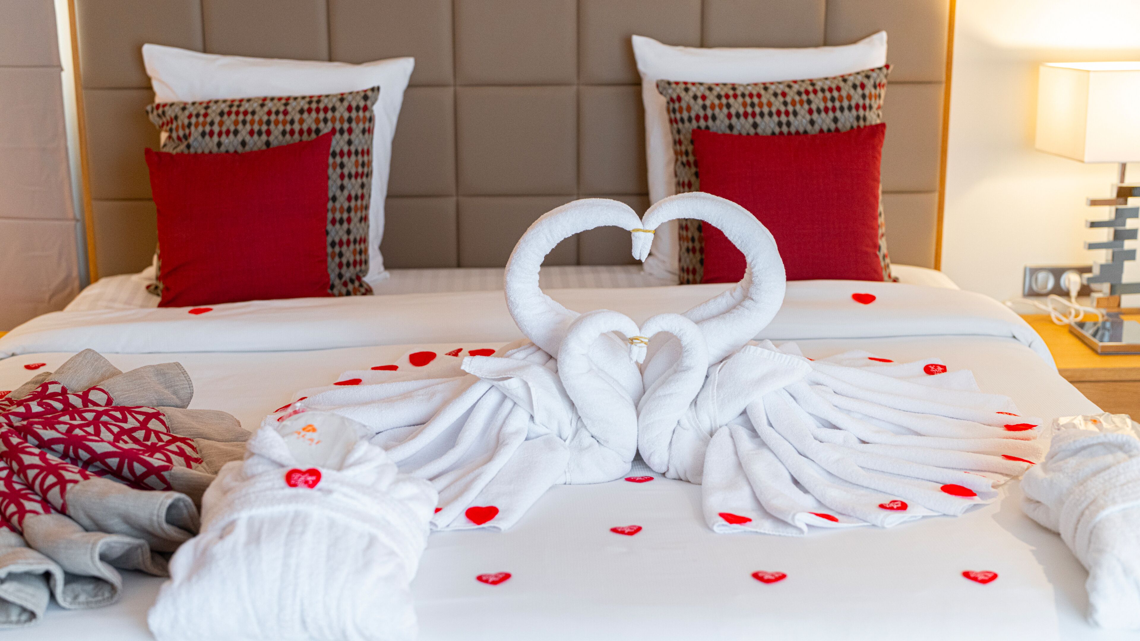 A hotel room with a bed featuring a heart-shaped towel arrangement and red heart-shaped decorations.