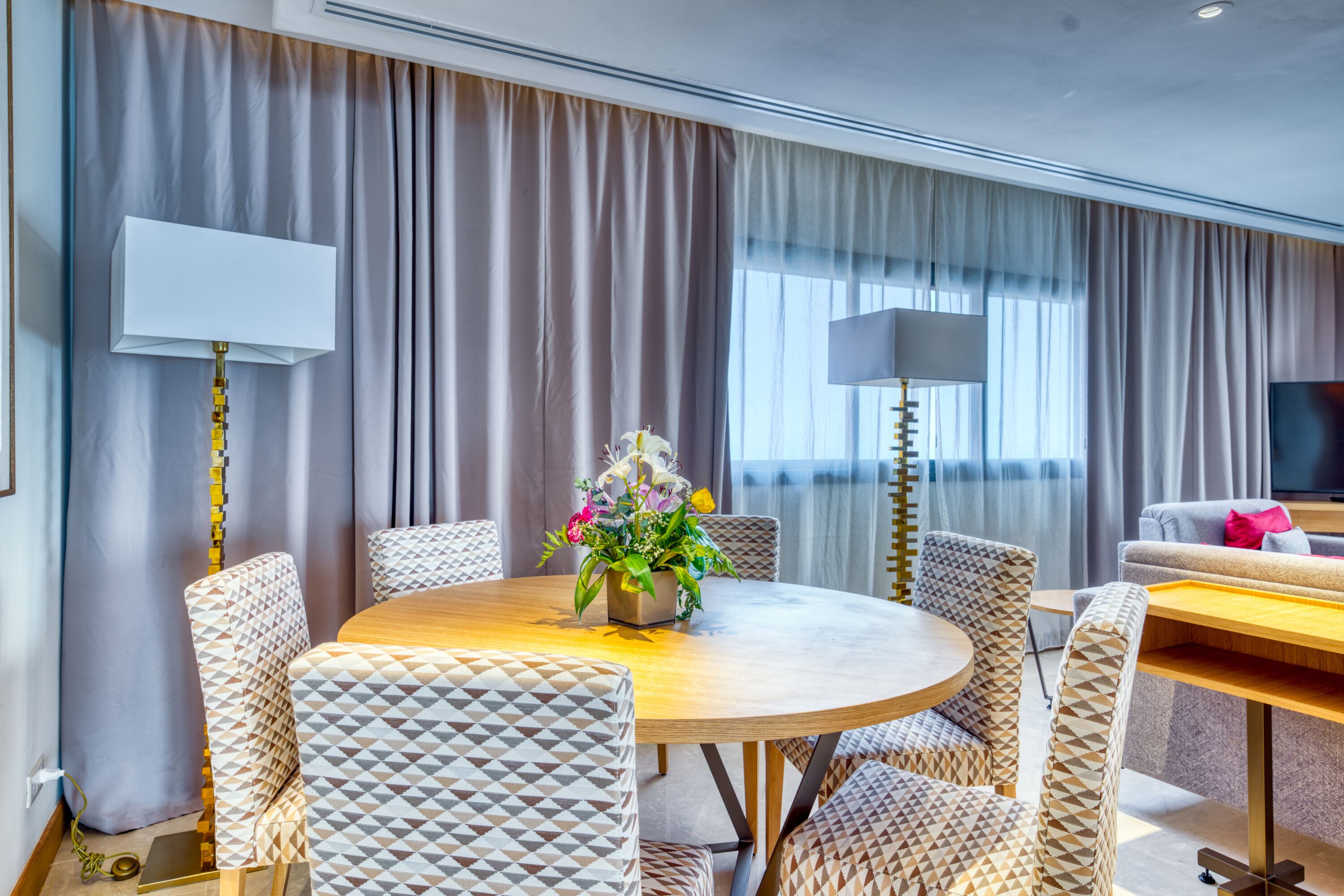 A hotel room with a round dining table, patterned chairs, a flower vase, a sofa, and a TV.