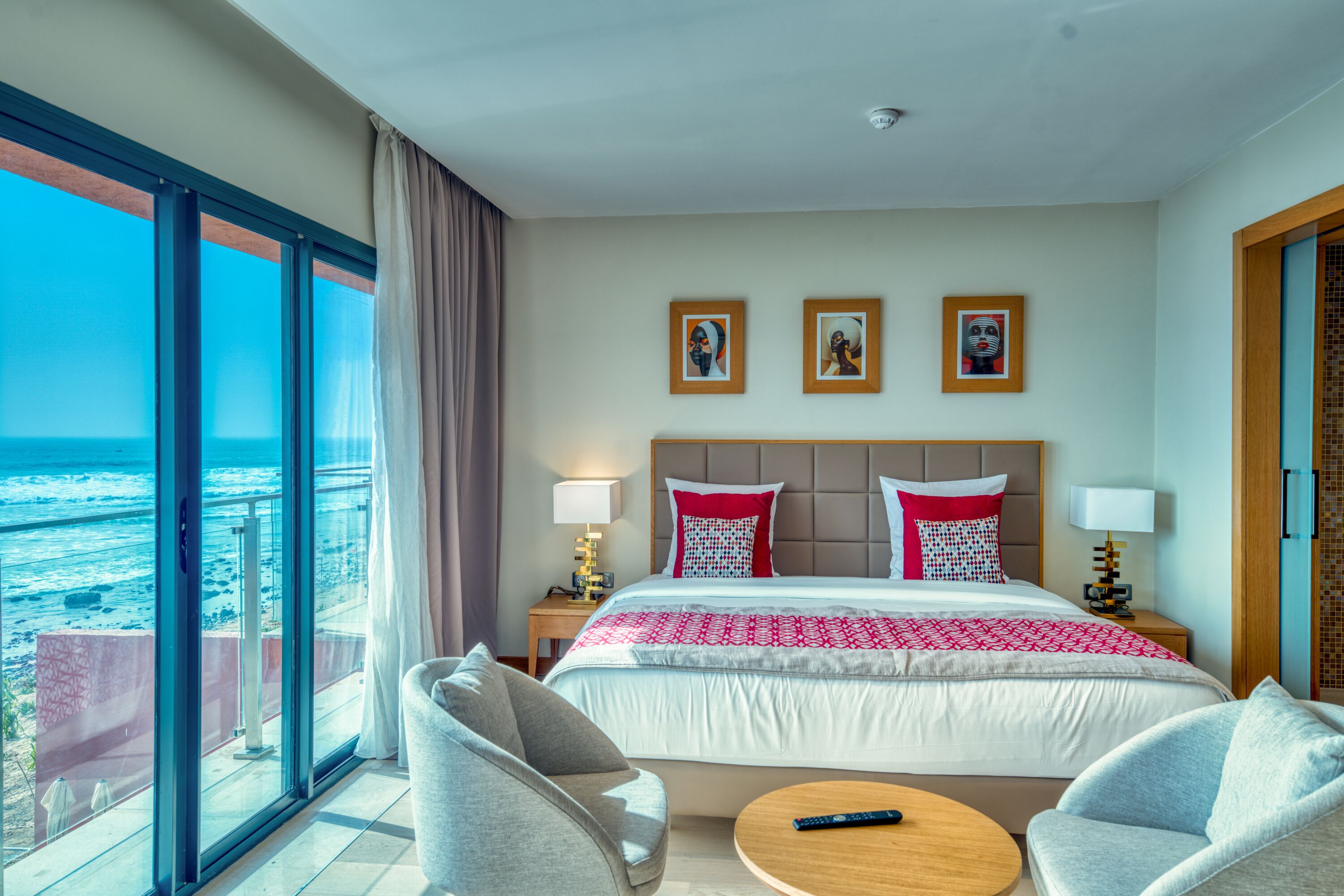 A hotel room with a large bed, two bedside lamps, a nightstand, a round coffee table, and a chair. There are three framed pictures on the wall above the bed.