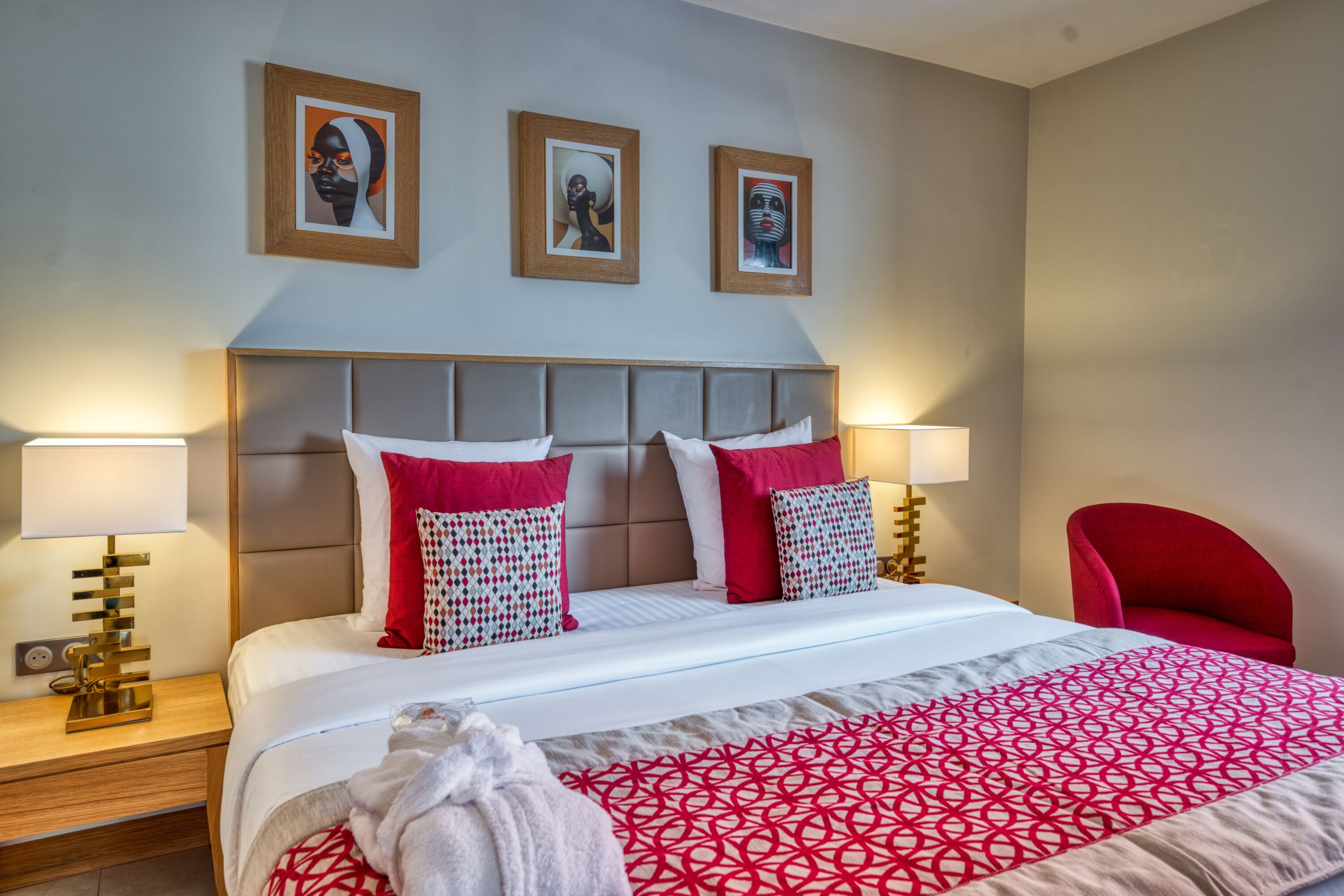 A hotel room with a bed, two bedside lamps, a red chair, and three framed pictures on the wall.