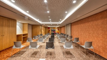 Een congreszaal met rijen stoelen, een houten paneelwand en een kamerbreed tapijt met dessin.