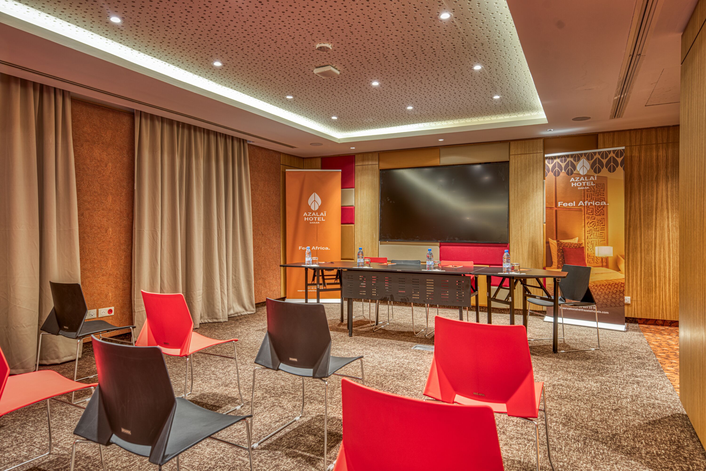 A conference room with a large screen, red chairs, a table with water bottles, and a banner for Azalai Hotel.