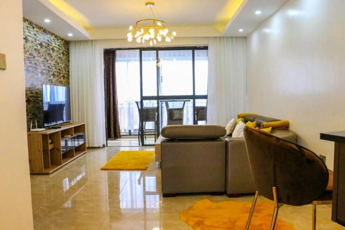 Una sala de estar moderna con un sofá, un televisor sobre un mueble de madera, una zona de comedor con una mesa y sillas, y un ventanal grande con cortinas.