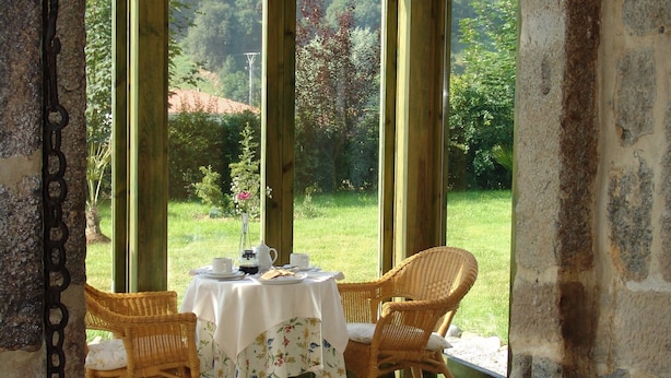 Ein Zimmer mit gedecktem Tisch für zwei Personen, Korbstühle, Blick auf einen Garten durch große Fenster und Steinwände.