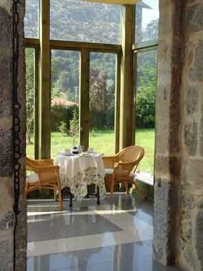 Ein Zimmer mit gedecktem Tisch für zwei Personen, Korbstühle, Blick auf einen Garten durch große Fenster und Steinwände.
