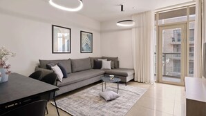 A modern living room with a grey sofa, a round coffee table, and a dining table with chairs. There are two framed artworks on the wall, a large window with curtains, and a balcony visible through the window. - Luxurious 1BDR Terrace Parking in City Central (Jerusalem)
