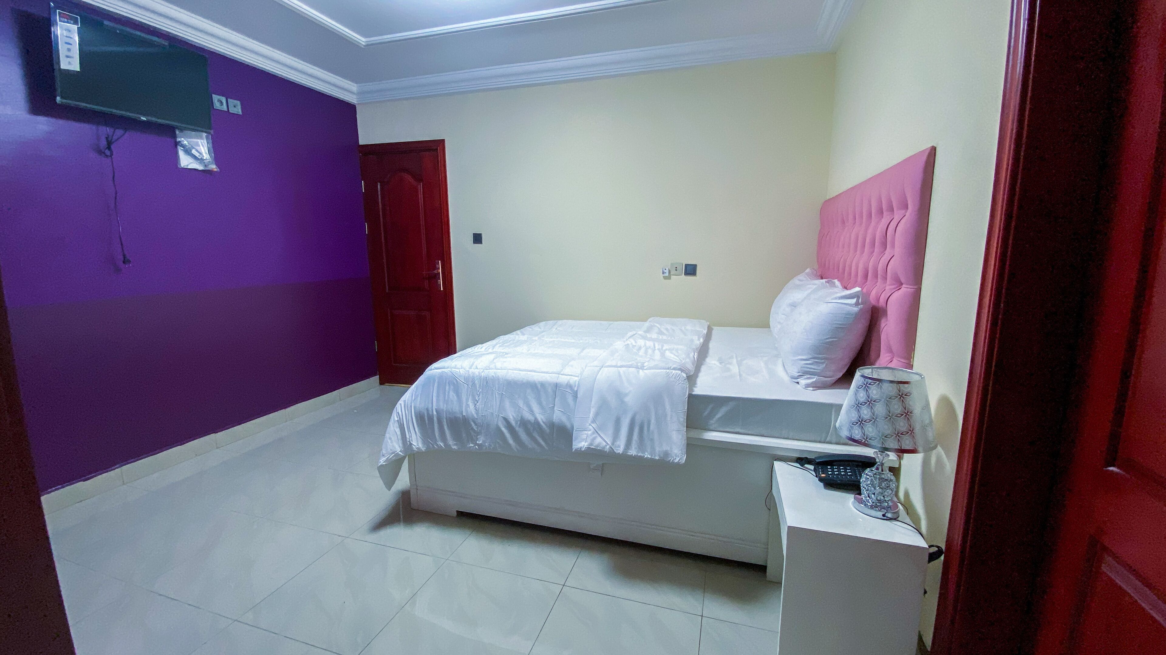 A bedroom with a bed, a desk, a chair, a TV mounted on the wall, and a door.