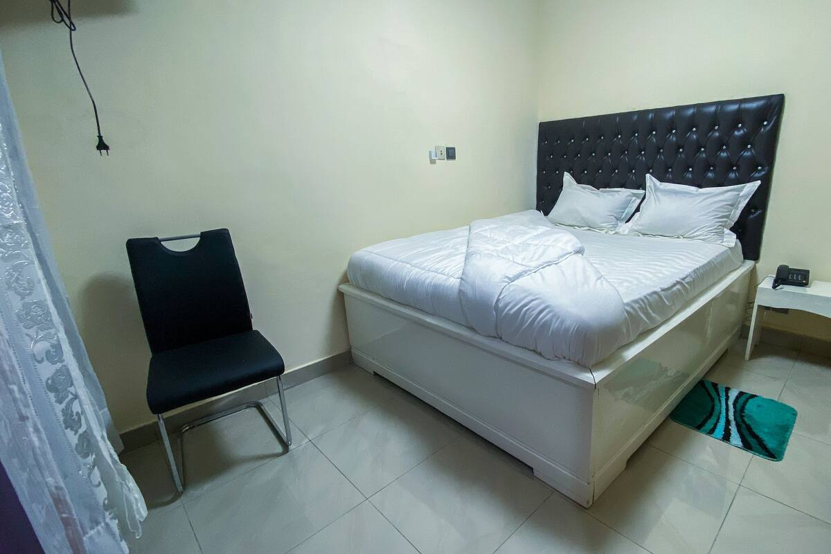 Un dormitorio con cama, una silla, una mesita de noche con teléfono y un enchufe en la pared.