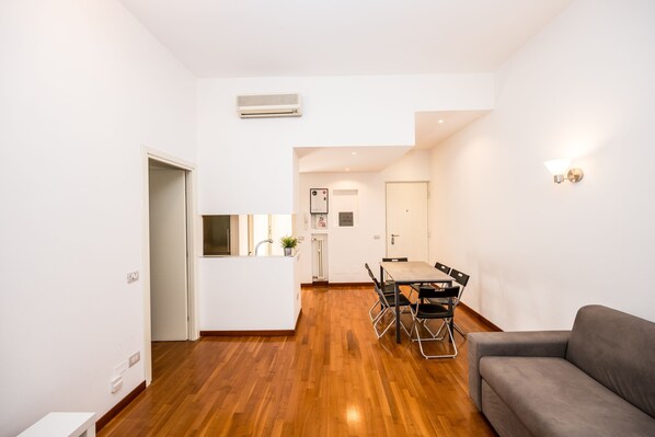 A modern living space with a wooden floor, a dining area with a table and chairs, a gray sofa, and a white wall with a light fixture.