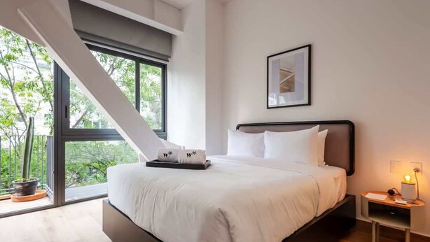 A modern bedroom with a large bed, a nightstand with a lamp, and a window with a view of greenery. - w* | Stunning loft in Cuauhtemoc (Ciudad de México)