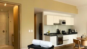 Una habitación de hotel moderna con cama, una mesita de noche con una toalla, una kitchenette con estufa y microondas, y una mesa de comedor de madera con sillas.