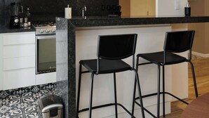Una cucina moderna con mobili bianchi, un piano di lavoro nero e un forno integrato. Sono presenti due sgabelli neri e un cestino.