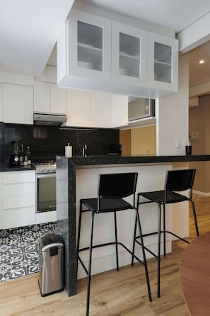 A modern kitchen with white cabinets, a black countertop, and a built-in oven. There are two black bar stools and a trash can. - w* | Vibrant 2BR with Balcony in Condesa (Ciudad de México)