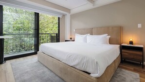 Ein modernes Schlafzimmer mit einem großen Bett, einem Nachttisch mit Lampe und Blick auf Grün durch eine Balkontür.