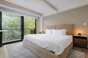 A modern bedroom with a large bed, a nightstand with a lamp, and a view of greenery through a balcony door.