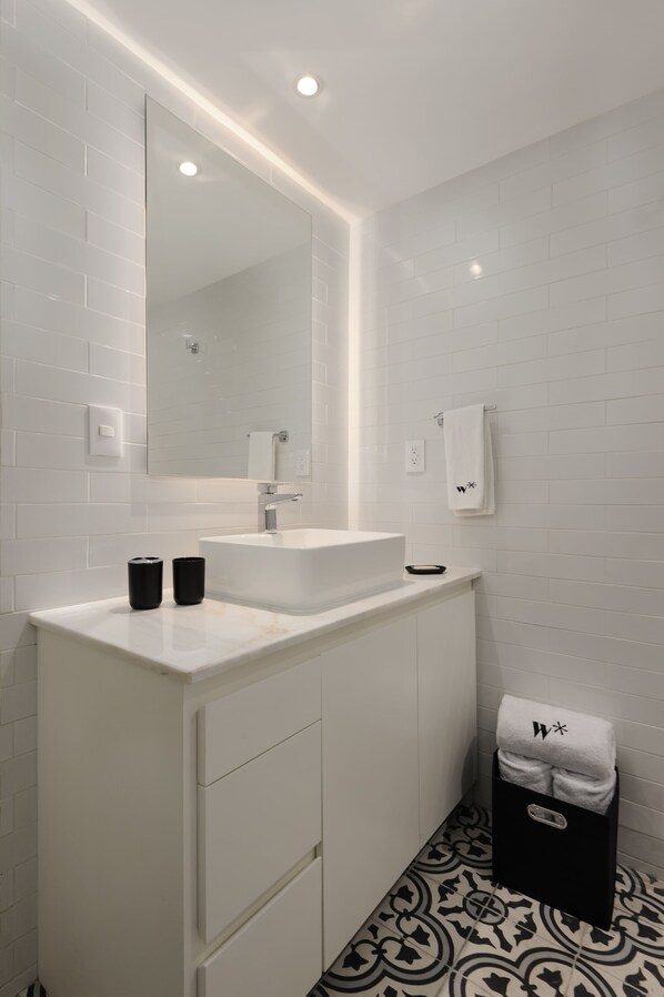 A modern bathroom with a white sink, mirror, and tiled walls. There is a towel rack with a towel and a black trash bin.