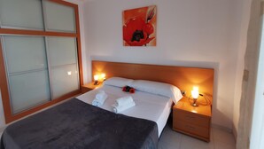 A hotel room with a bed, two bedside tables, a nightstand, a wall-mounted painting, and a window with blinds. - Beachside Apartment with Free WiFi, Terraces & Near Shops and Restaurants (Platja d'Aro)