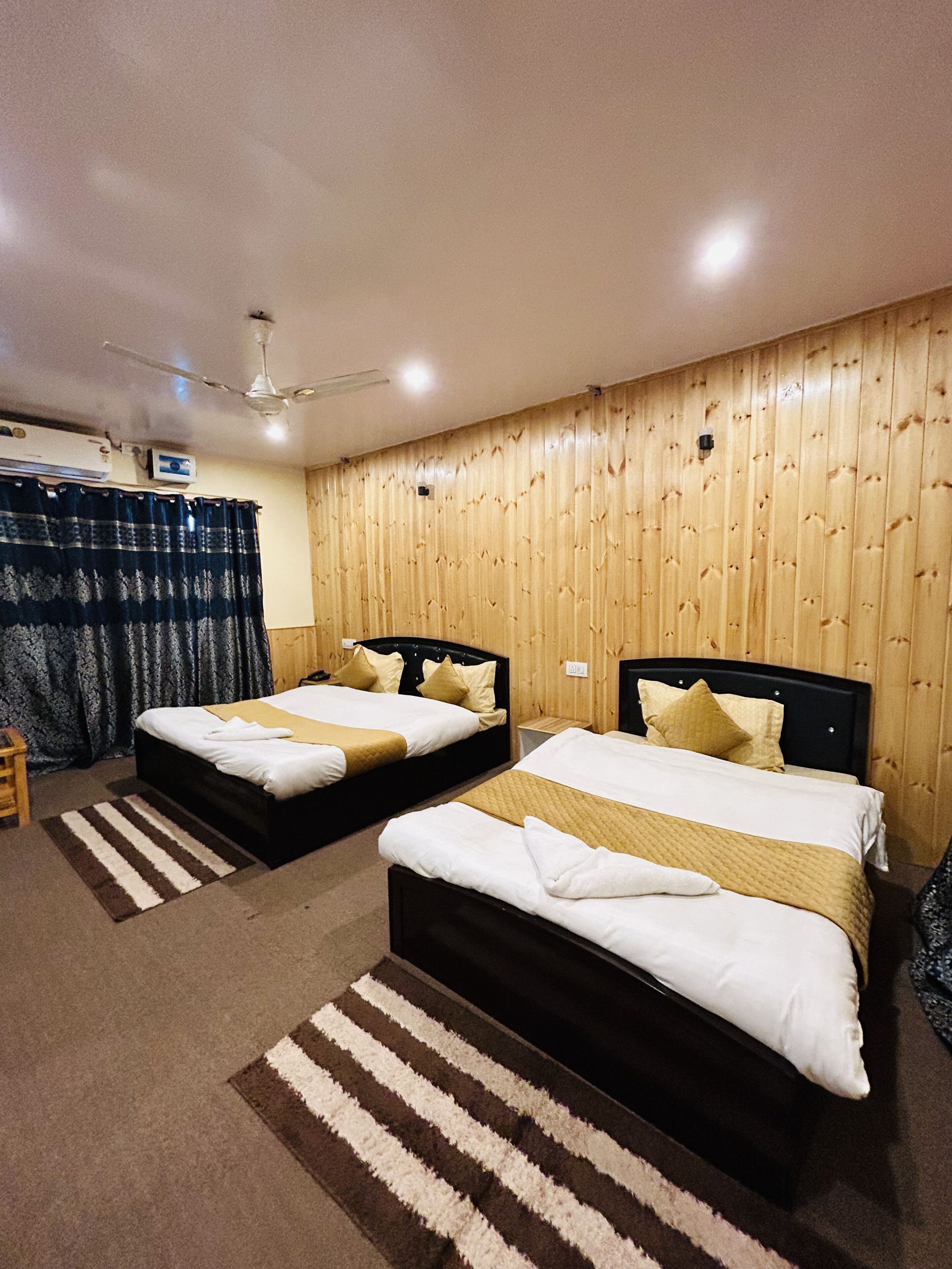 A hotel room with two beds, wooden panel walls, a ceiling fan, and a window with curtains.