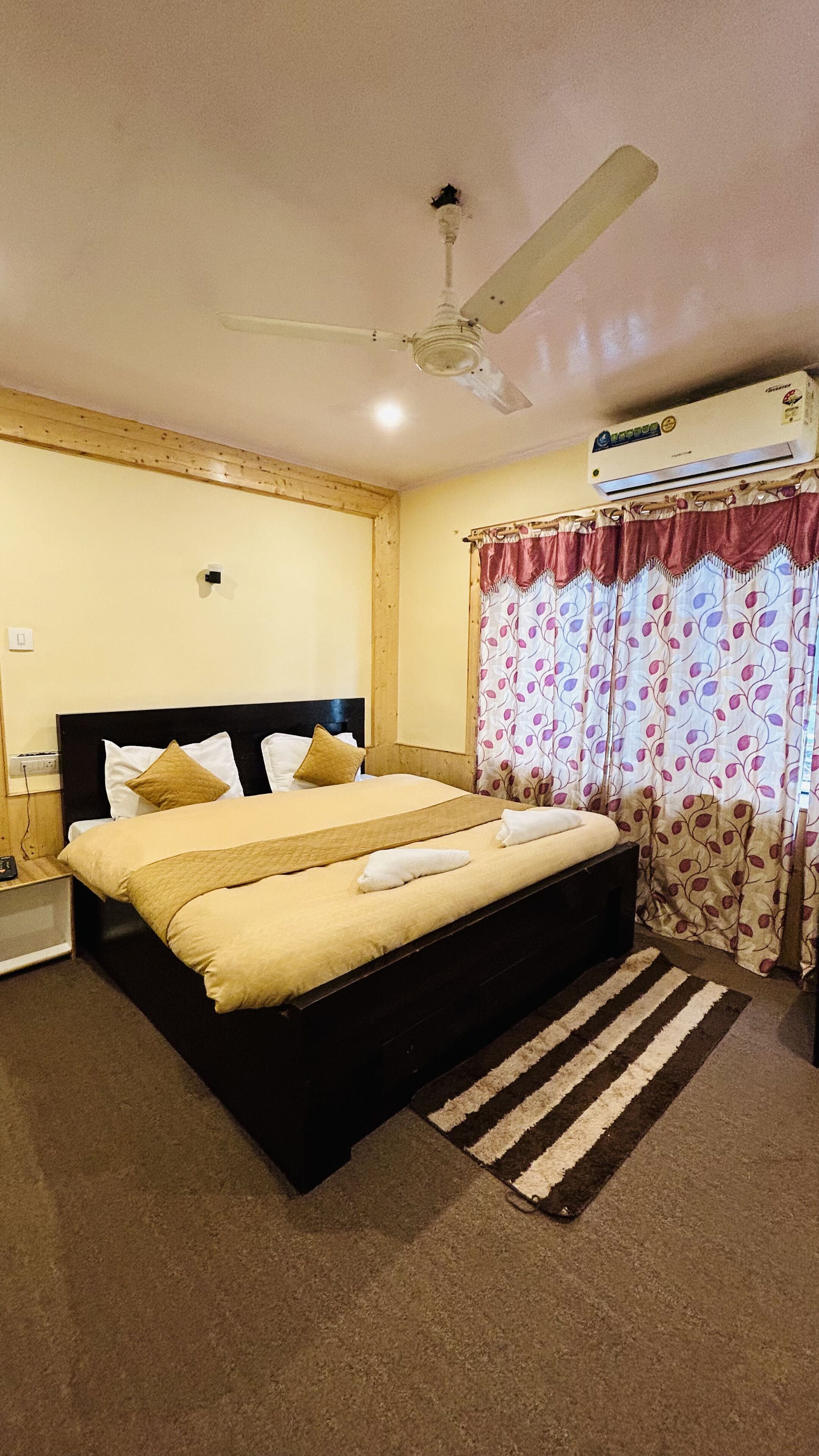A bedroom with a bed, a ceiling fan, an air conditioner, a window with curtains, and a carpeted floor.