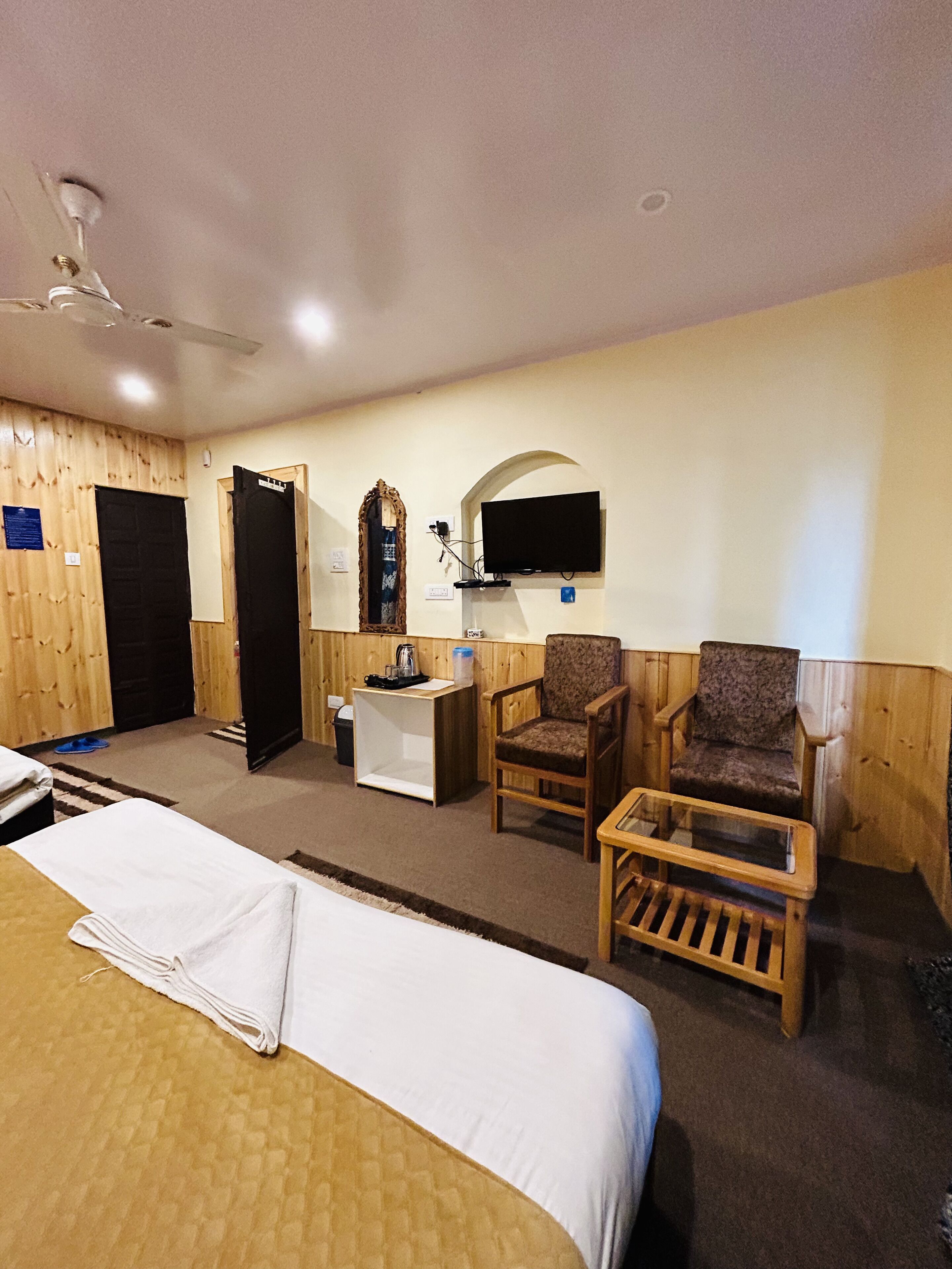A hotel room with a bed, two chairs, a small table, a television mounted on the wall, and a ceiling fan.
