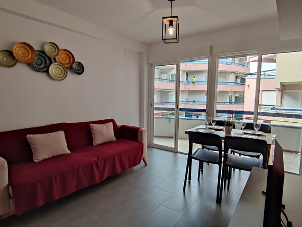 Un salon avec un canapé rouge, une table à manger avec des chaises et un balcon offrant une vue sur les immeubles.