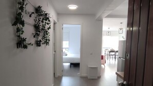 Un pasillo que conduce a un dormitorio con una cama, una mesita y sillas. Hay una puerta a la derecha y una planta a la izquierda.