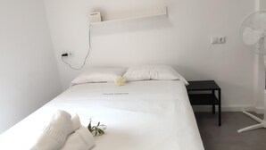 Habitación de hotel minimalista con una cama bien tendida, una mesita de noche, un estante en la pared y un ventilador de pie.