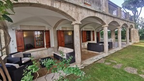 Uma área de estar coberta ao ar livre, com colunas de pedra e persianas de madeira, cercada por um gramado bem cuidado e uma grande árvore.