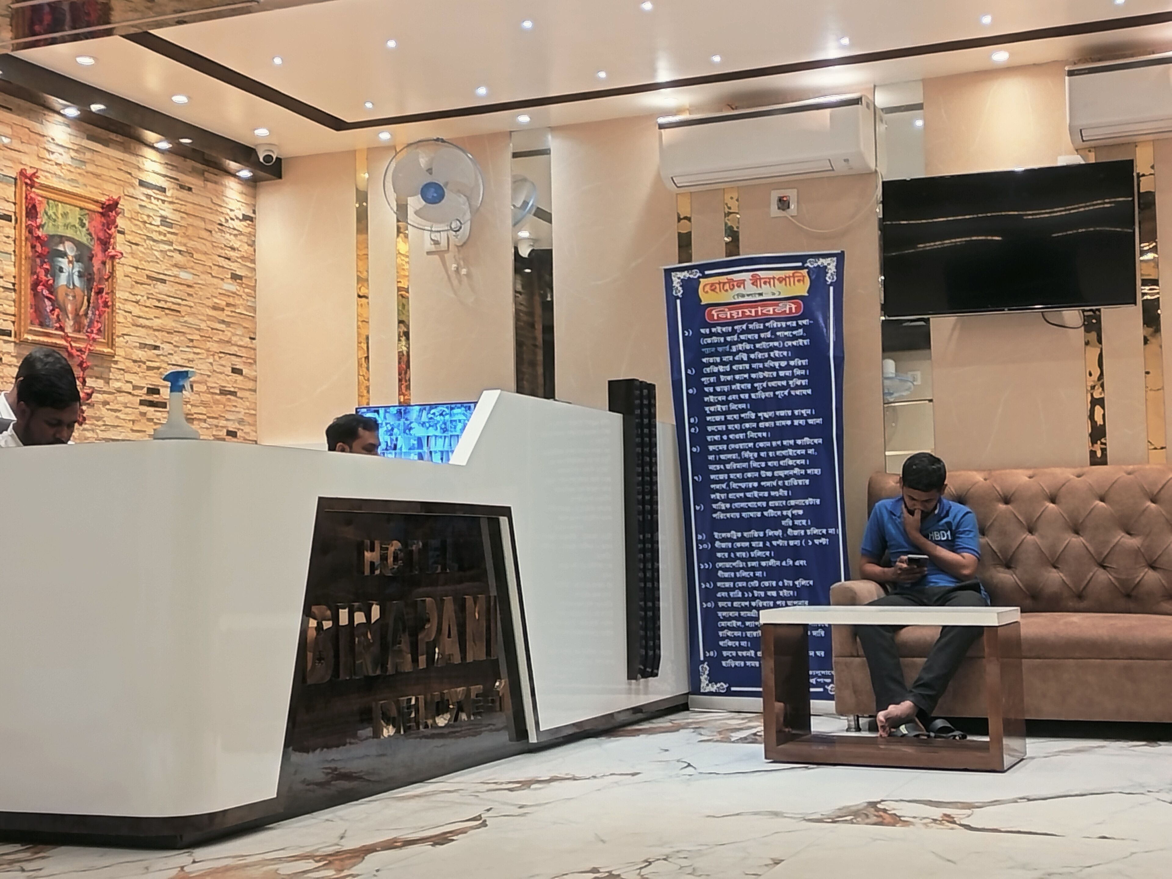 Een hotel receptie met een receptiebalie, een groot verticaal bord met tekst, een aan de muur bevestigde televisie en een zithoek met een bank.