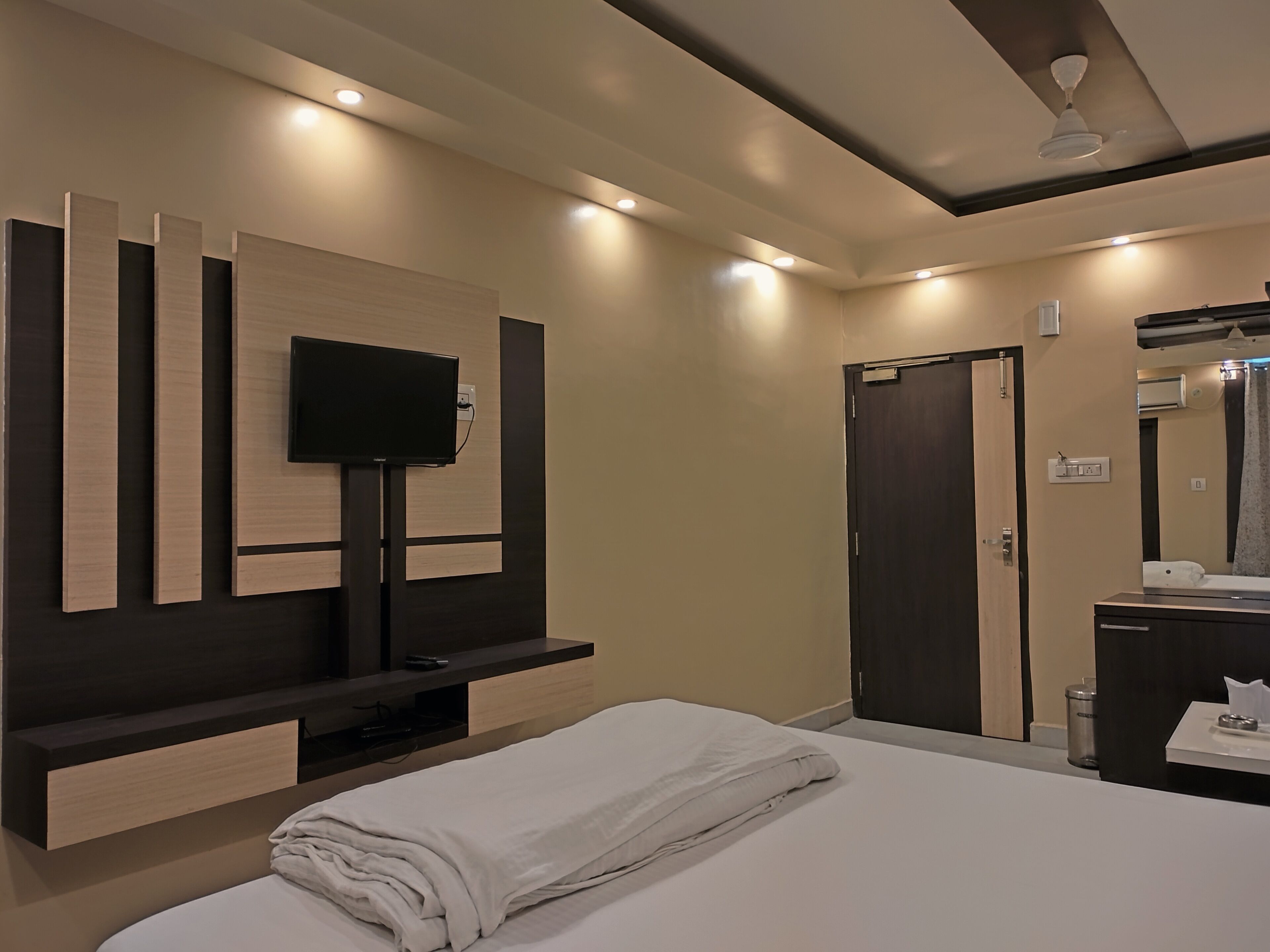 A modern hotel room with a bed, a TV mounted on the wall, a dark door, and a bathroom with a mirror and sink.