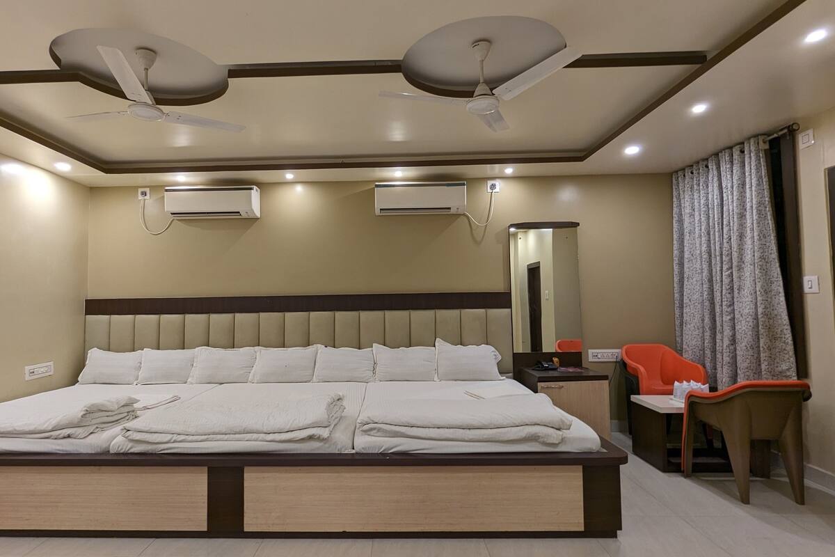 Una habitació d'hotel moderna amb un llit gran, ventiladors de sostre, aires condicionadors, una taula de nit, una cadira, un mirall i una porta.