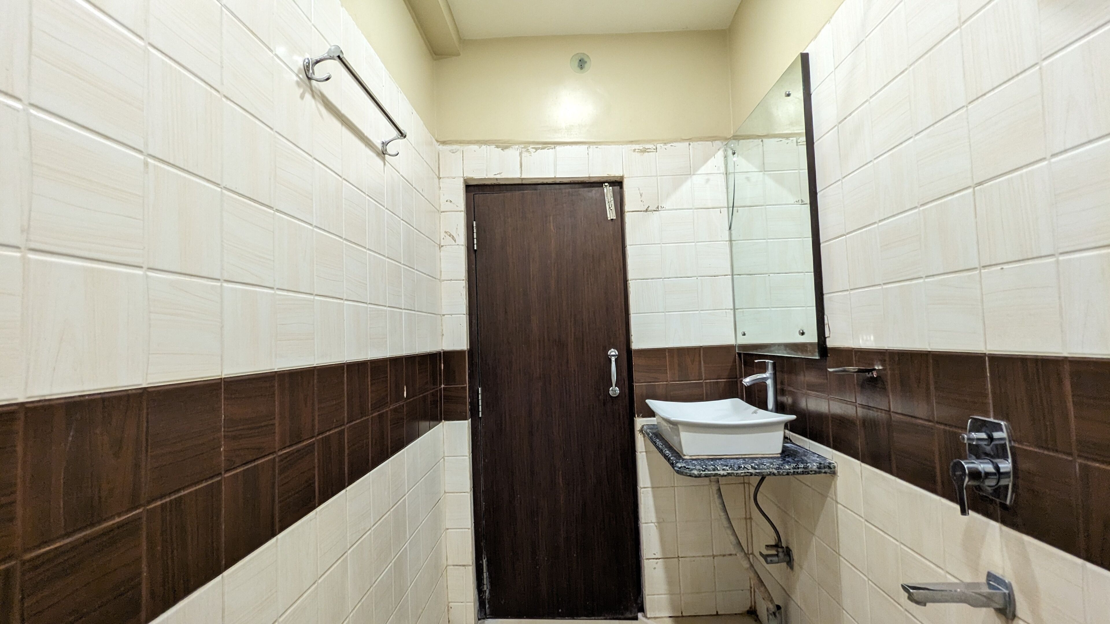 Un baño pequeño con azulejos blancos y marrones, una puerta de madera, un lavamanos con espejo y un perchero para toallas.