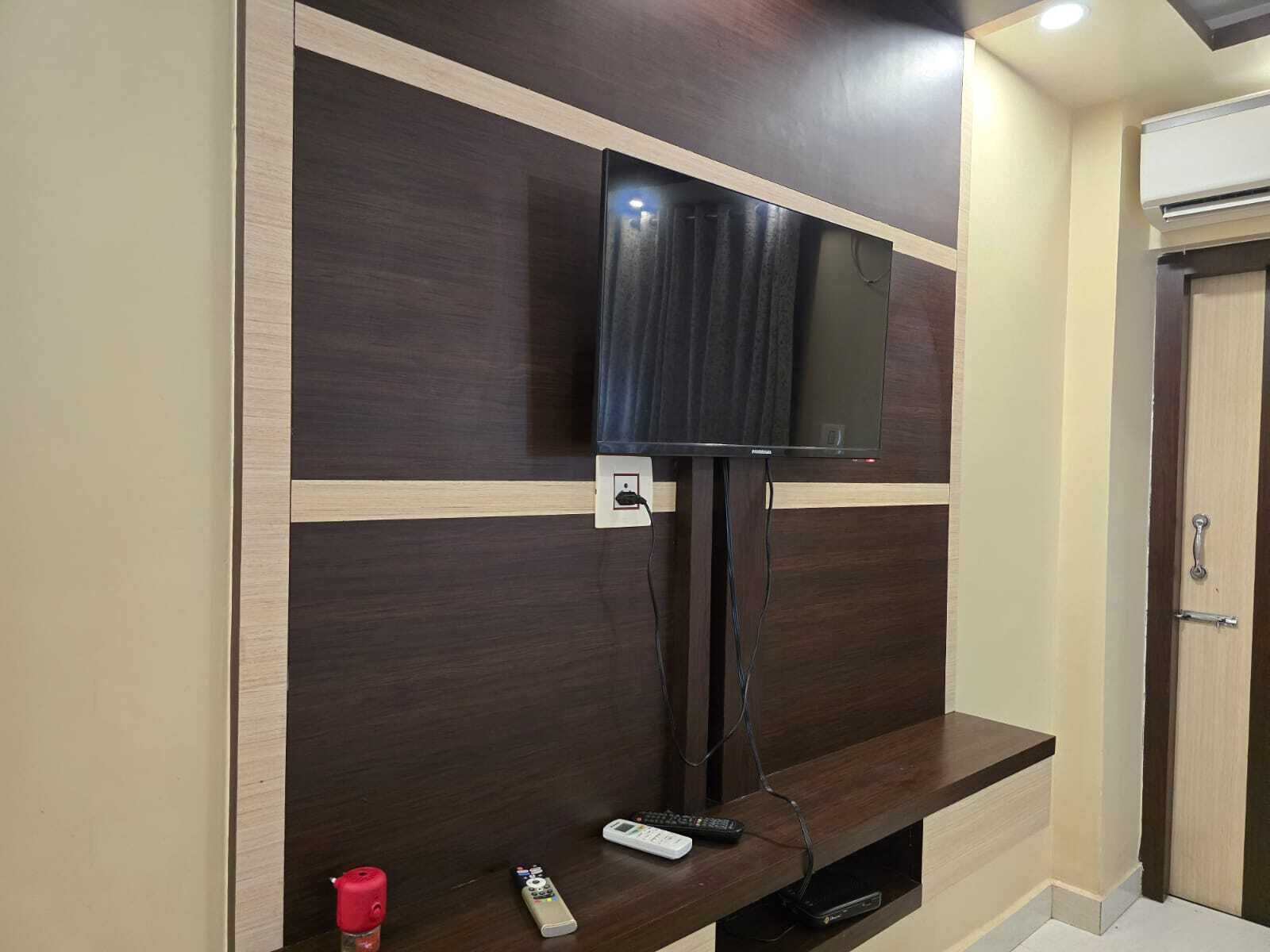 A room with a flat-screen TV mounted on a dark wood panel wall, a small console table with a remote control, and a door with an air conditioning unit above it.