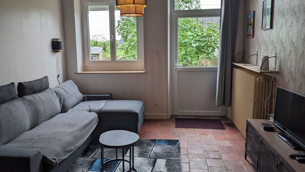 Ein gemütliches Wohnzimmer mit einer Couch, einem kleinen runden Tisch, einem Fernseher und einem Fenster mit Blick ins Grüne.