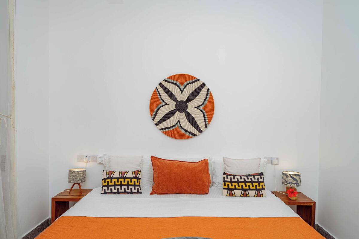 Una cama bien hecha con una colcha naranja, almohadas blancas y una almohada naranja decorativa. Un tapiz redondo con un diseño geométrico está centrado sobre la cama.