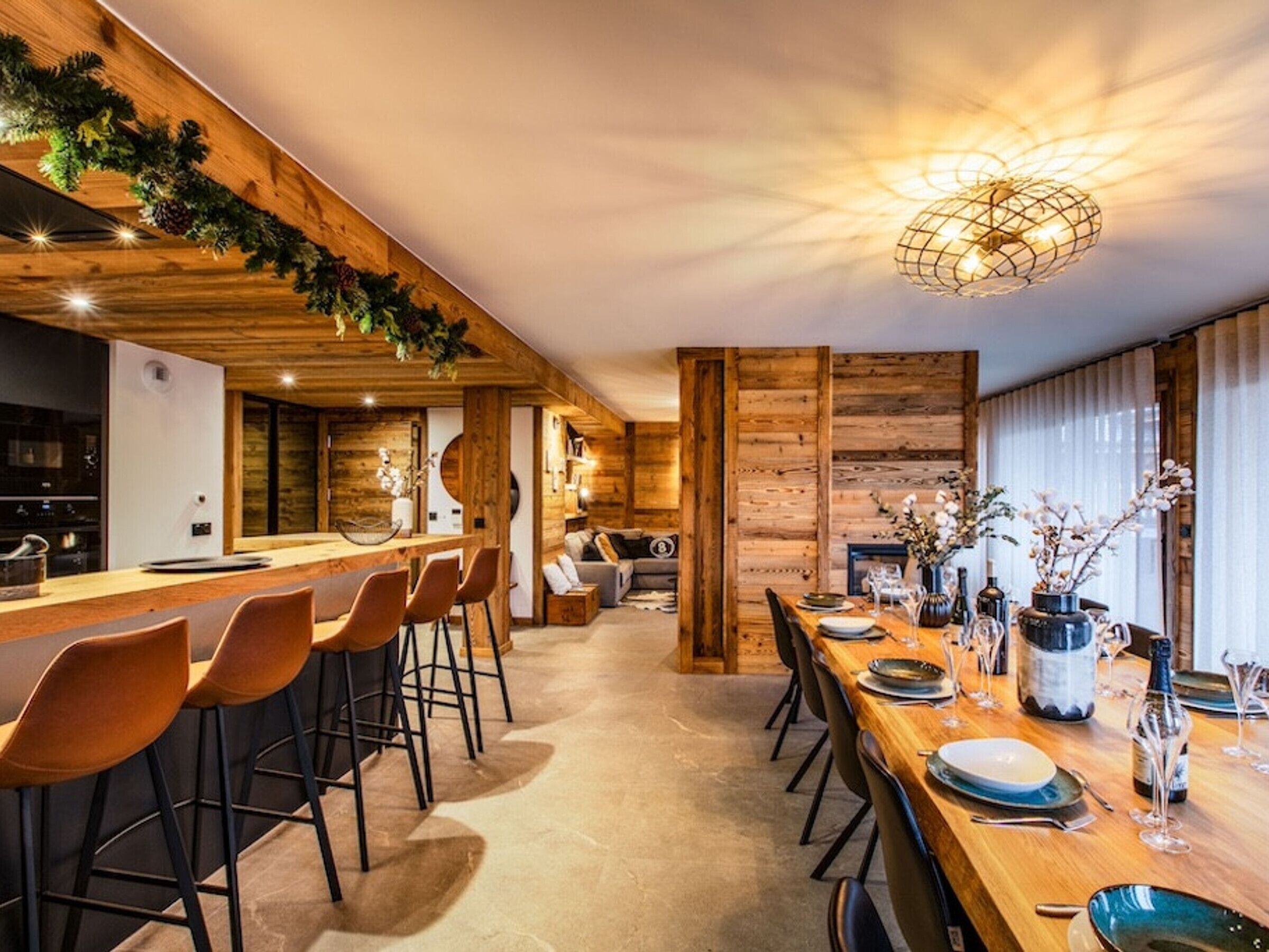 A modern kitchen with wooden accents, a bar area with stools, and a dining table set for six with wine glasses and bottles.