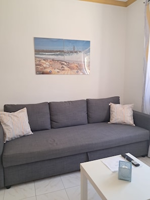 A grey sofa with striped pillows, a white coffee table with a remote control, and a beach scene painting on the wall. - Largo Frente - Familiar & Acolhedor (Quarteira)