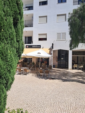 A café with outdoor seating, a white building with balconies, and a cobblestone courtyard. - Largo Frente - Familiar & Acolhedor (Quarteira)