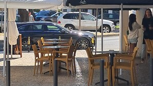Un espacio al aire libre con mesas y sillas, una zona techada y vista a la calle con coches estacionados y árboles.