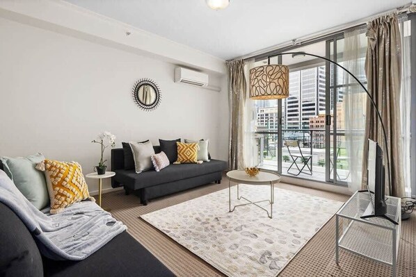 A modern living room with a sofa, a coffee table, and a TV stand. There is a large window with curtains and a view of buildings outside.