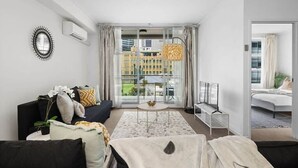 A modern living room with a black sofa, a coffee table, and a TV. There is a large window with a view of buildings outside. - A Modern Apt for 6 Right Next to Darling Harbour (Sydney)