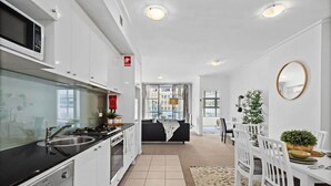 A modern kitchen with white cabinets, a black countertop, and a built-in oven. There is a dining area with white chairs and a table, and a living room with a black sofa and a window. - A Modern Apt for 6 Right Next to Darling Harbour (Sydney)
