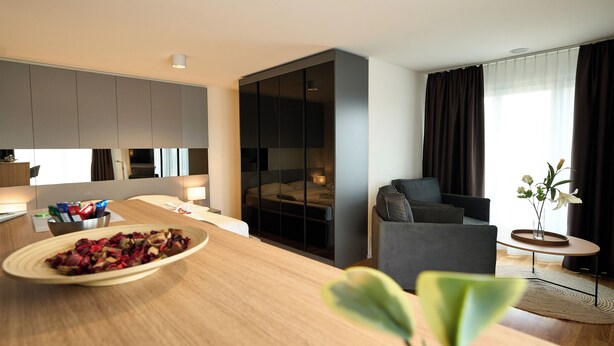 A modern living room with a sofa, a dining table with a bowl of fruit, and a large window with curtains. - Airport Apartments Zürich (Opfikon)