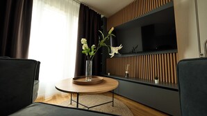 A modern living room with a round wooden table, two dark sofas, a TV mounted on a wooden panel wall, and a vase with flowers on the table. - Airport Apartments Zürich (Opfikon)