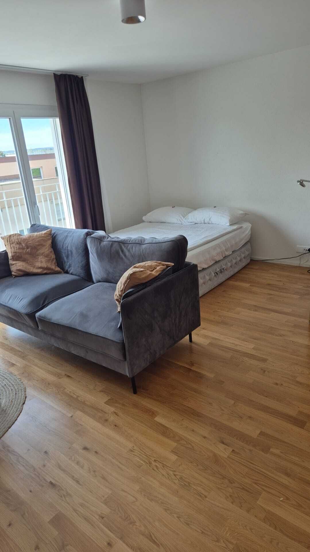 A modern living room with a grey sofa, a mattress, and a wooden floor.