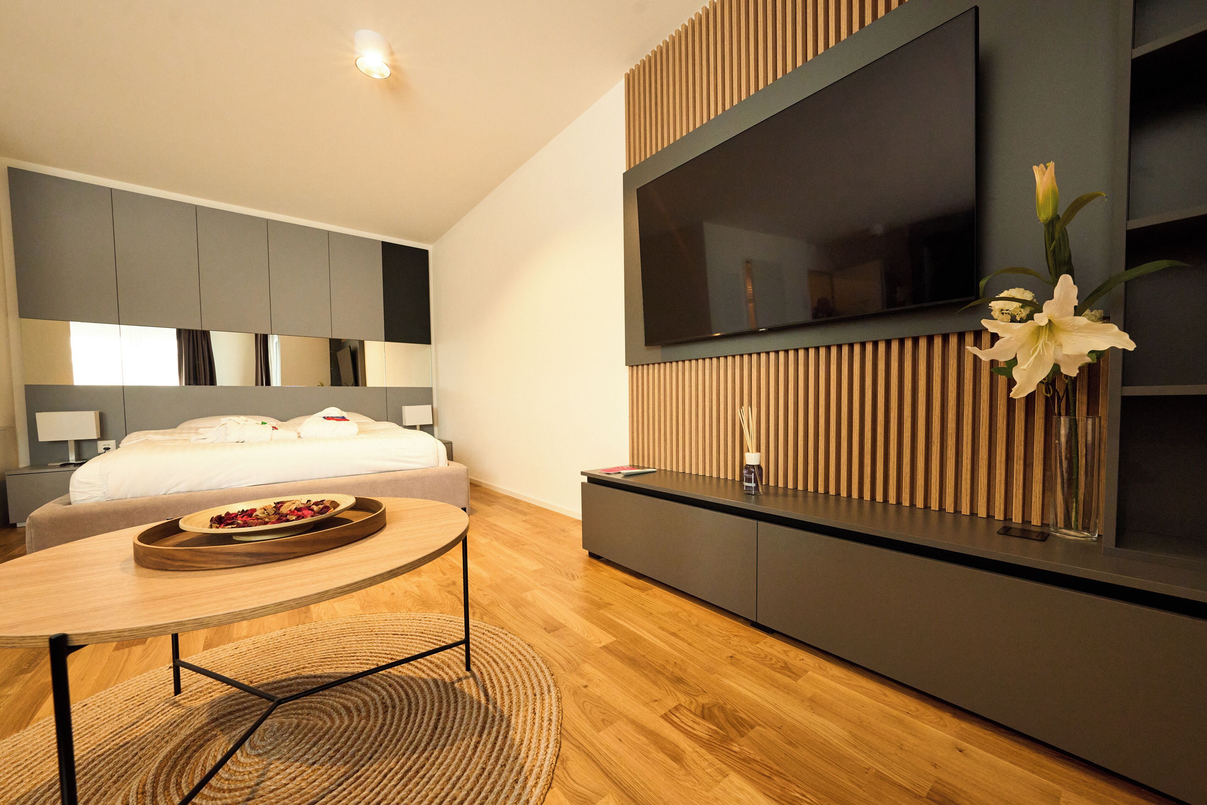 Ein modernes Hotelzimmer mit Bett, einem an einer Holzplatte montierten Fernseher, einem kleinen Tisch mit Tablett und einer Vase mit Blumen.