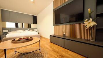 Ein modernes Hotelzimmer mit Bett, einem an einer Holzplatte montierten Fernseher, einem kleinen Tisch mit Tablett und einer Vase mit Blumen.