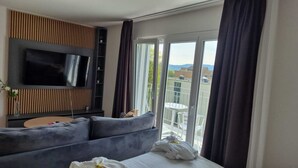 A modern hotel room with a large flat-screen TV mounted on a wooden panel, a grey sofa, a small round table, and a balcony with a glass door. - Airport Apartments Zürich (Opfikon)