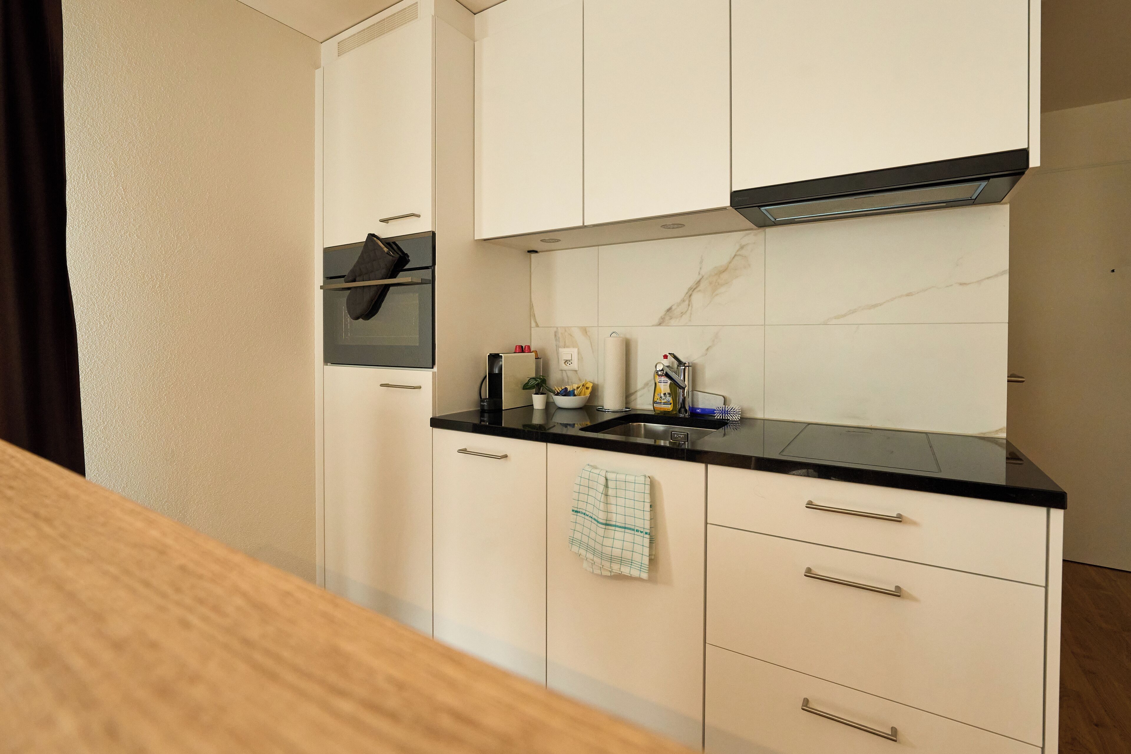 A small kitchen with white cabinets, a black countertop, and a microwave. There is a sink with a faucet, a towel hanging on the cabinet, and a few kitchen items on the counter.